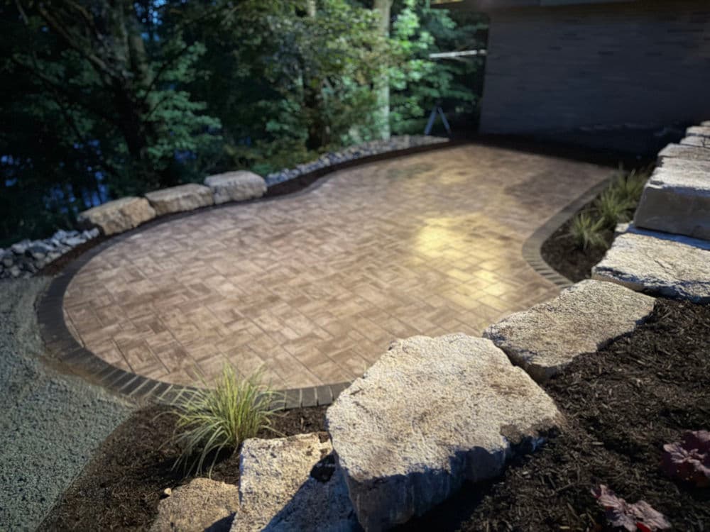 Patterned stone patio surrounded by landscaping and natural stone border in wooded setting.